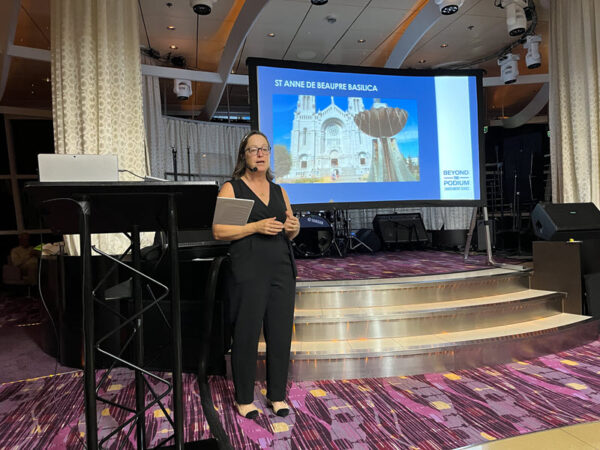 A staff member on a cruise, using a headworn microphone. A PowerPoint presentation is in the back