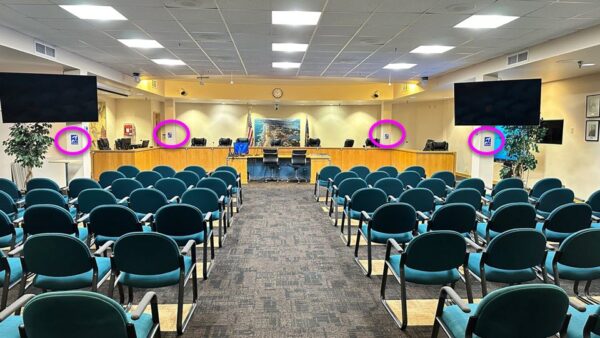 empty city council room looking forward to bench. 2 assistive listening signs on each side are circled